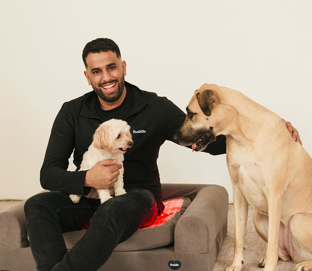 A smiling man sitting with a small white dog and a large tan dog on a Buddie red light therapy bed, showing the bed's size versatility